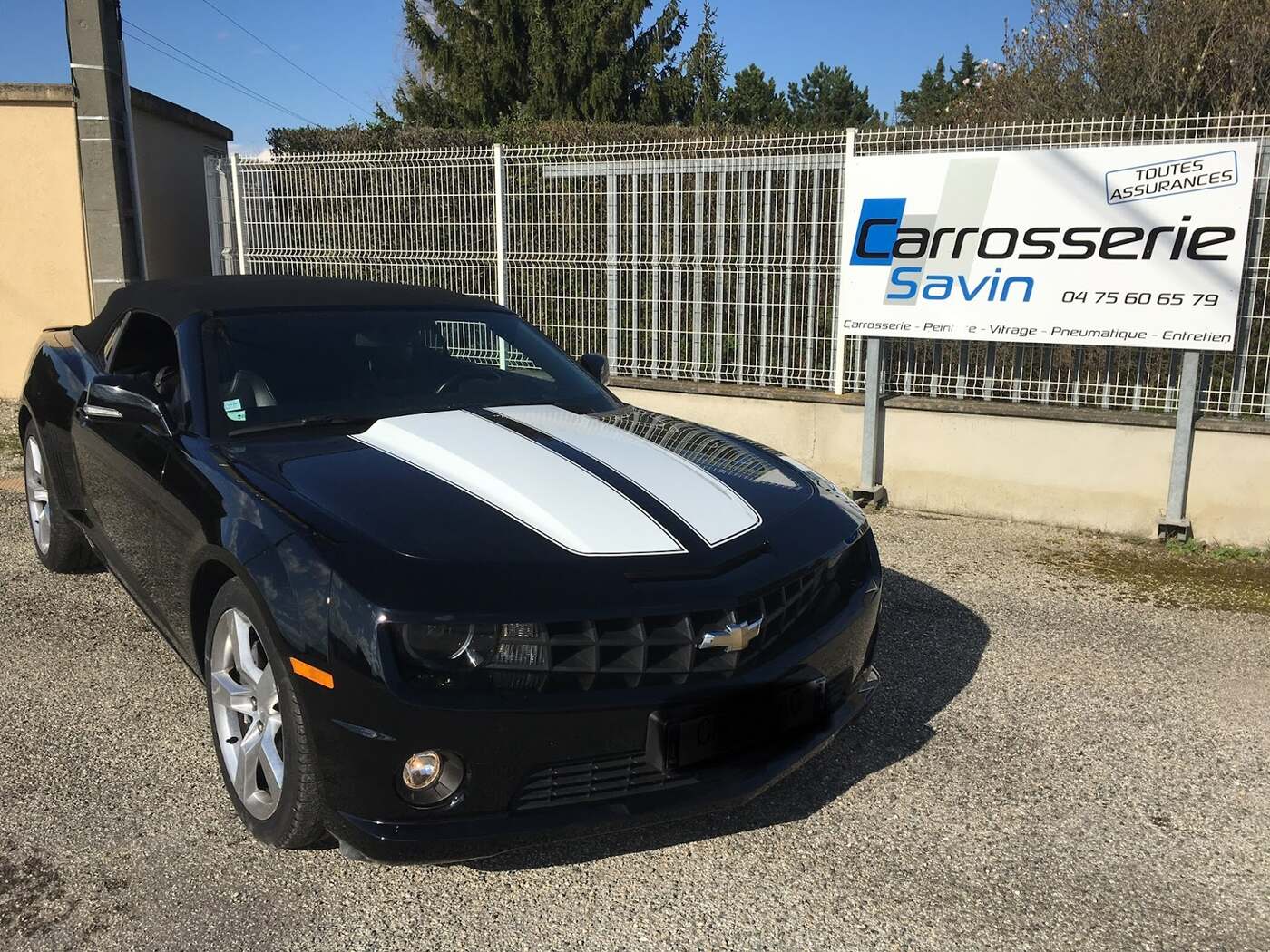 Une Camaro cabriolet noire à deux bandes blanches sur le capot, garée sur le gravier devant le panneau Carrosserie Savin fixé à la clôture de l'atelier.