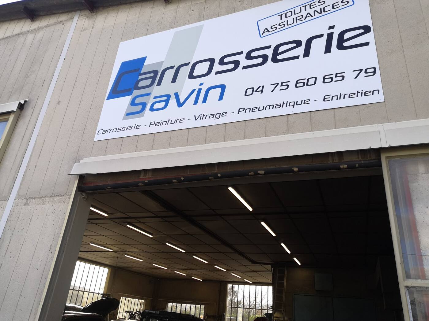 Façade en béton de l'atelier avec l'enseigne Carrosserie Savin au-dessus de la grande porte ouverte sur l'atelier éclairé.